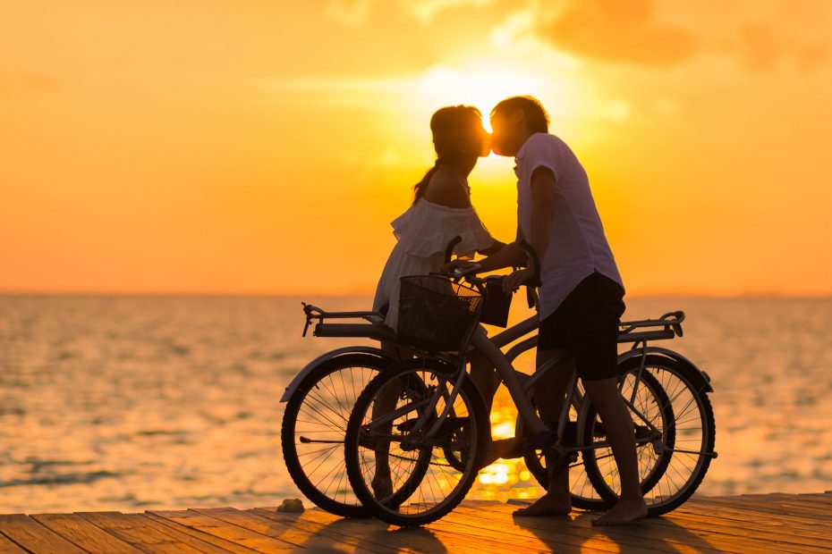 photography of man wearing white t shirt kissing a woman while holding bicycle on river dock during sunset